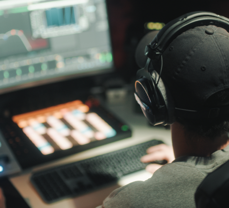  A view of the back of a person wearing a baseball cap and overear headphones, sitting in front of a screen with an open music program.