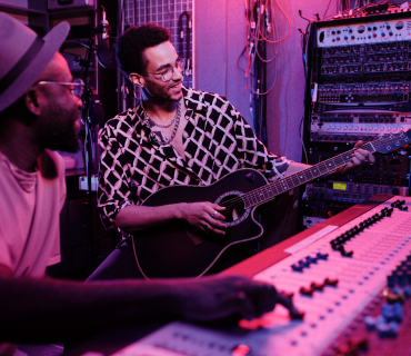 Two men: one operates a mixing console, the other plays guitar.