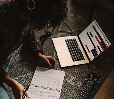 Creating your own song Person sitting on the floor with headphones reading in a notebook next to open laptop